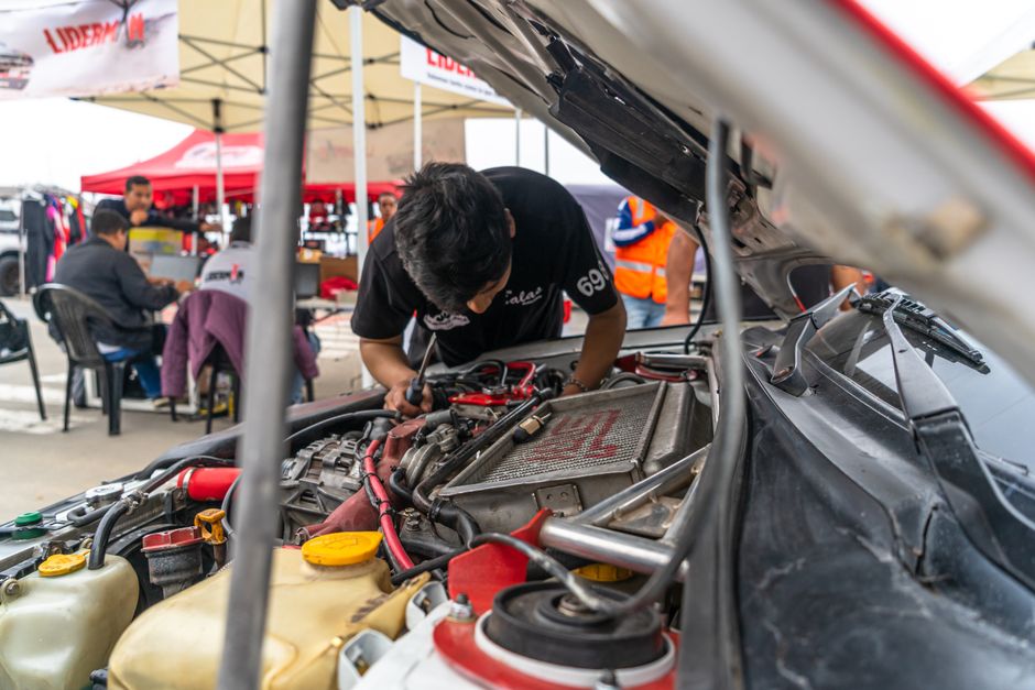 Mechanic inspecting a car engine outdoors at an automotive workshop. Team members in the background.
